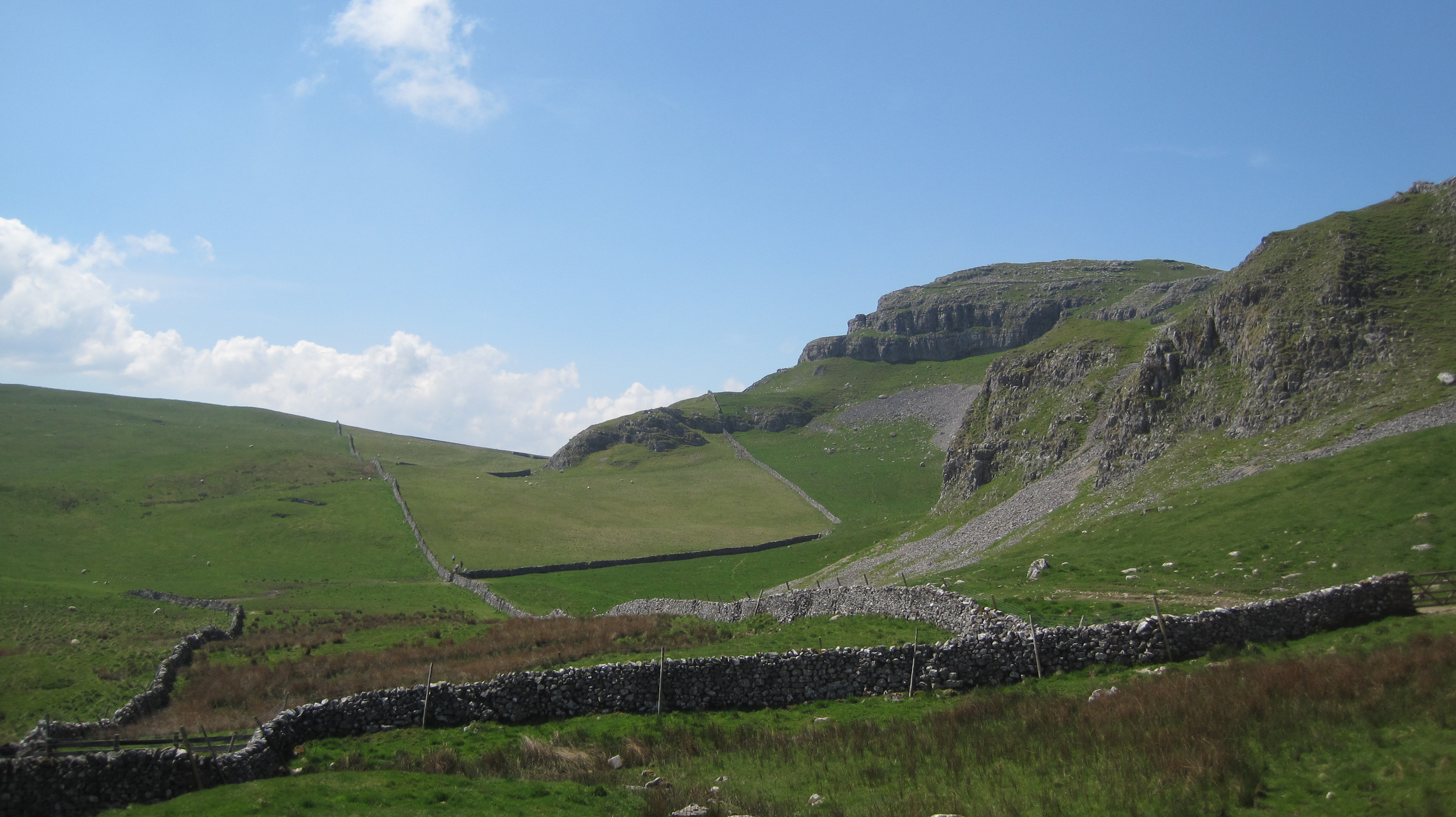 The British countryside with a steep grassy slope and a drystone wall against a hazy blue sky.